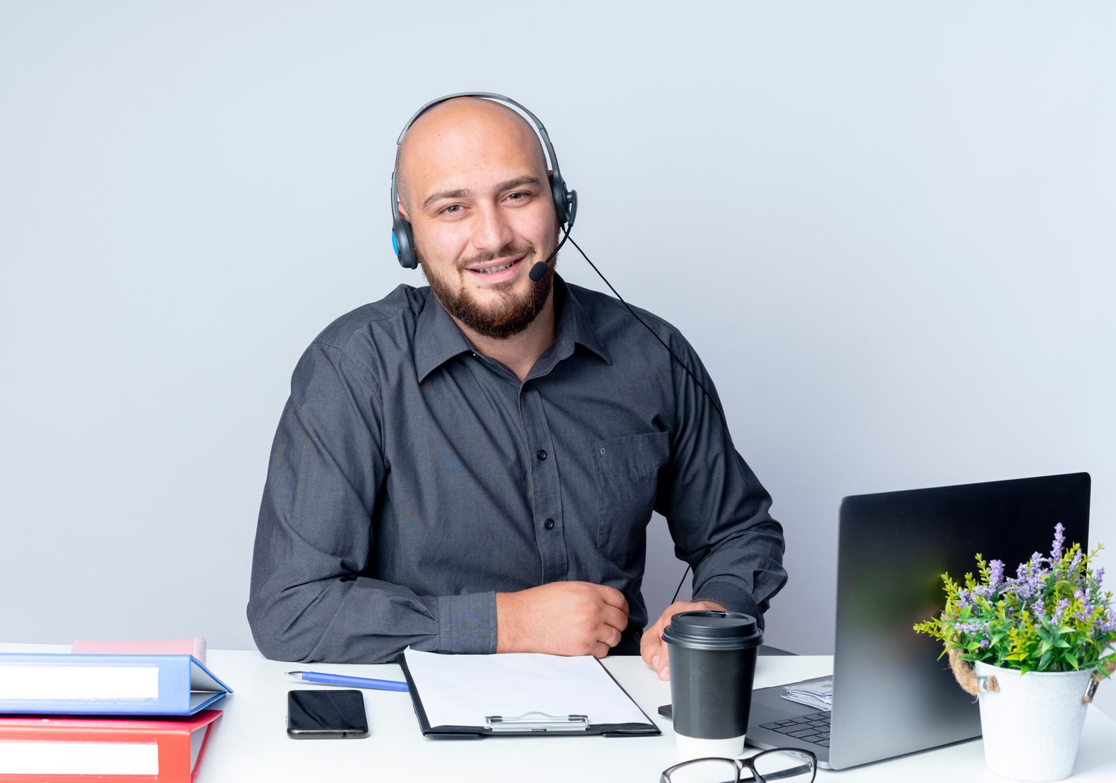 smiling young bald call center man wearing headset sitting at desk with work tools putting hands on desk isolated on white background